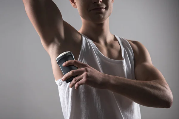 Vista recortada de un joven con camisa blanca sin mangas aplicando desodorante en la axila aislada en gris - foto de stock