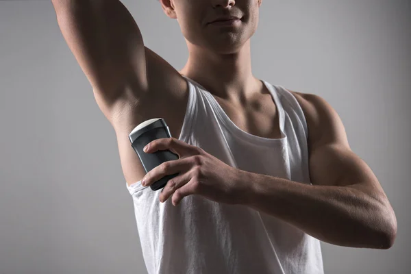 Vue recadrée du jeune homme en chemise blanche sans manches appliquant du déodorant sur les aisselles isolées sur gris — Photo de stock