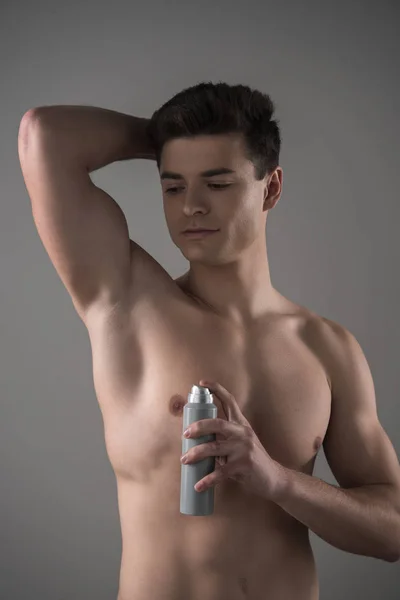 Giovane uomo senza camicia che tiene deodorante vicino alla ascella isolato su grigio — Foto stock