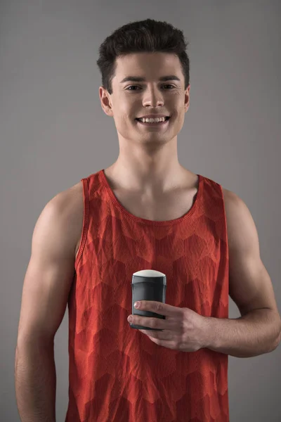 Young man in red sleeveless shirt holding deodorant while smiling at camera isolated on grey — Stock Photo