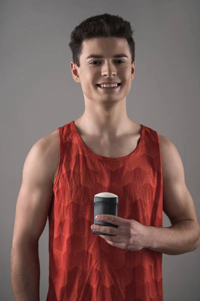 Young man in red sleeveless shirt holding deodorant while smiling at camera isolated on grey — Stock Photo