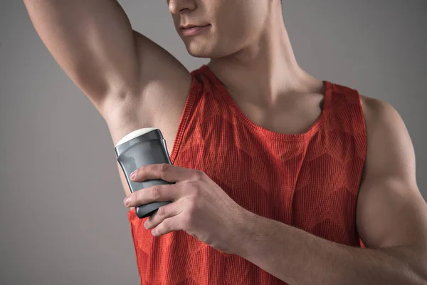 Vista recortada del joven con camisa roja sin mangas aplicando desodorante en la axila aislada en gris - foto de stock