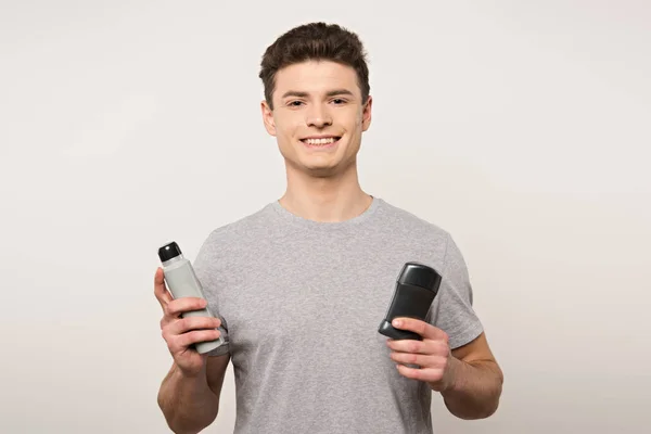 Young man in grey t-shirt holding deodorants and smiling at camera isolated on grey — Stock Photo
