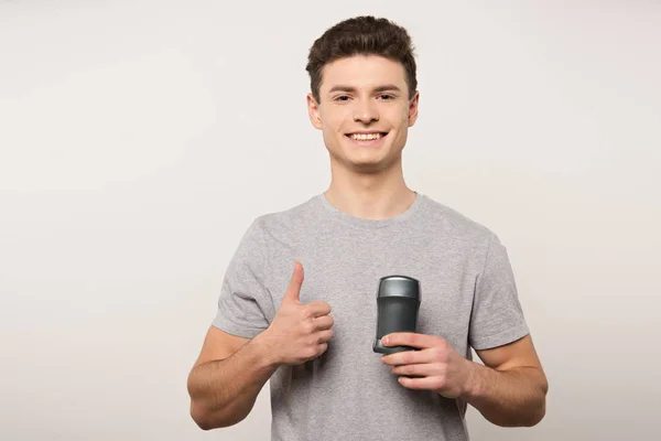 Smiling man in grey t-shirt holding deodorant and showing thumb up isolated on grey — Stock Photo