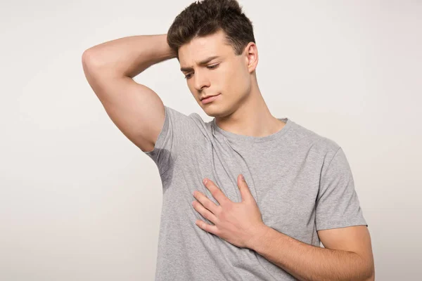 Displeased man in grey t-shirt with sweaty underarm holding hand of chest isolated on grey — Stock Photo