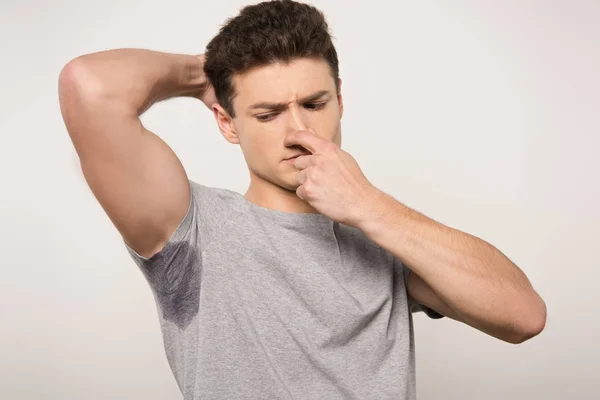 Displeased man in grey t-shirt with sweaty underarm plugging nose with hand isolated on grey — Stock Photo