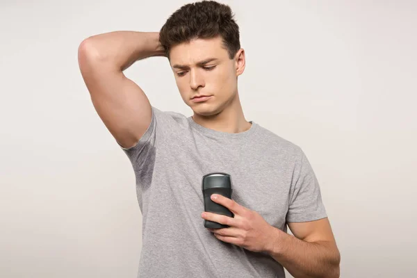 Homem descontente em camiseta cinza com suor na axila segurando desodorizante isolado em cinza — Fotografia de Stock