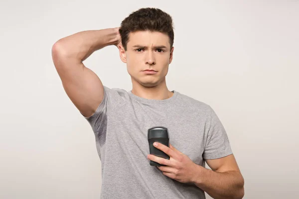 Homme mécontent en t-shirt gris avec les aisselles moites tenant déodorant isolé sur gris — Photo de stock