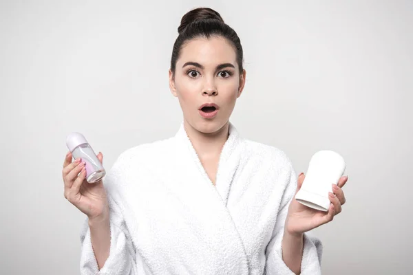 Surprised woman in bathrobe holding deodorants while looking at camera isolated on grey — Stock Photo