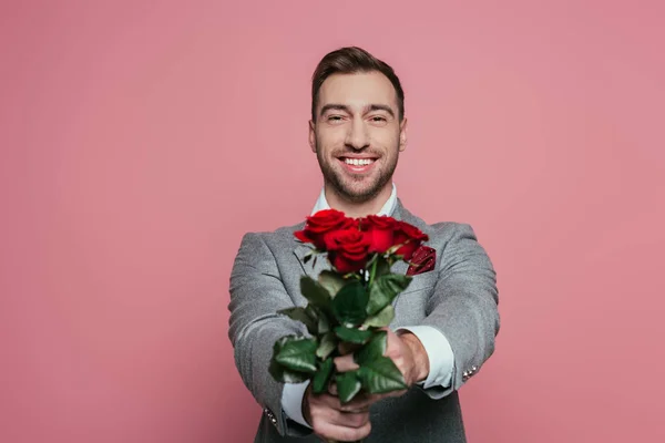 Apuesto hombre alegre en traje sosteniendo ramo de rosas rojas, aislado en rosa - foto de stock