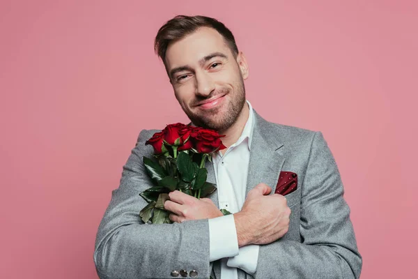 Красивий усміхнений чоловік у костюмі, що тримає червоні троянди, ізольований на рожевому — Stock Photo