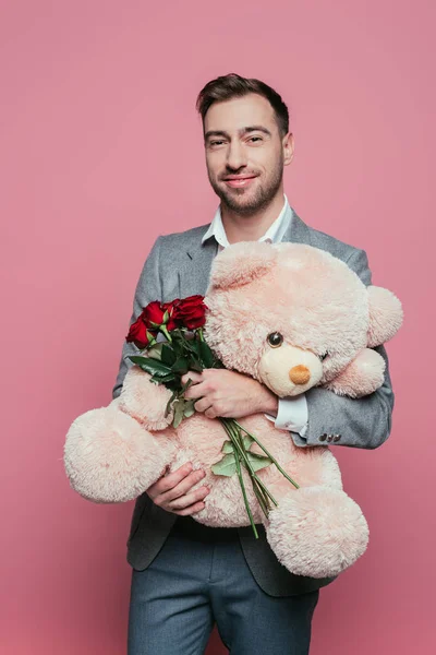 Apuesto hombre alegre sosteniendo osito de peluche y rosas rojas, aislado en rosa - foto de stock