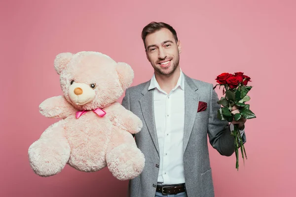 Homem sorridente segurando ursinho de pelúcia e rosas, isolado em rosa — Fotografia de Stock