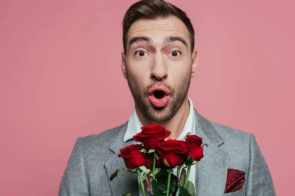 Здивований чоловік у костюмі тримає червоні троянди, ізольовані на рожевому — стокове фото