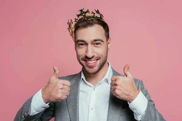 Красивый улыбающийся мужчина в короне и костюме показывает большие пальцы вверх, изолированные на розовый — стоковое фото