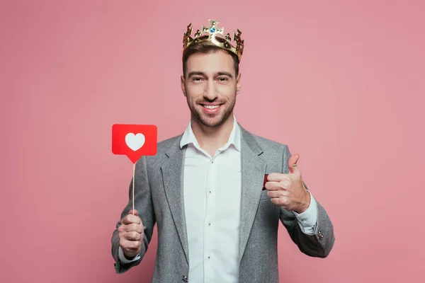 Hombre feliz en la corona mostrando el pulgar hacia arriba y la celebración de la tarjeta con el corazón para el día de San Valentín, aislado en rosa - foto de stock