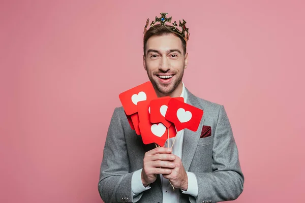Fröhlicher Mann in Krone mit Karten mit Herzen zum Valentinstag, isoliert auf rosa — Stockfoto