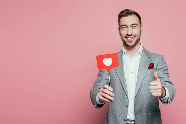 Hombre feliz mostrando el pulgar hacia arriba y la celebración de la tarjeta con el corazón para el día de San Valentín, aislado en rosa - foto de stock