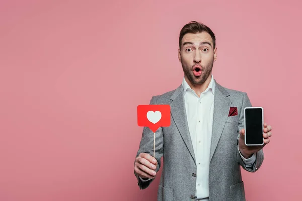 Homem surpreso segurando smartphone com tela em branco e cartão com coração para o dia dos namorados, isolado no rosa — Fotografia de Stock