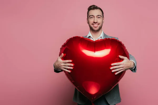 Hombre barbudo feliz sosteniendo globo rojo del corazón en el día de San Valentín, aislado en rosa - foto de stock
