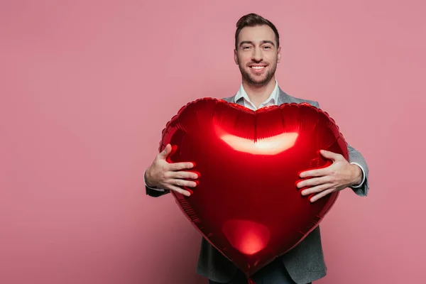 Felice uomo barbuto tenendo palloncino cuore rosso il giorno di San Valentino, isolato su rosa — Foto stock