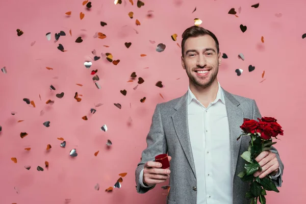 Uomo sorridente che tiene rose e proposta anello rosa con cuori coriandoli — Foto stock