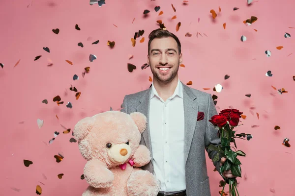 Улыбающийся мужчина в костюме держит плюшевого мишку и розы на День Святого Валентина, на розовом с конфетти — стоковое фото