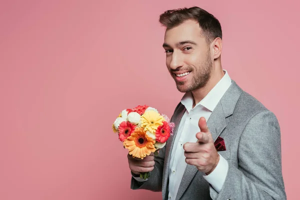 Homme heureux barbu en costume tenant des fleurs et pointant vers vous, isolé sur rose — Photo de stock