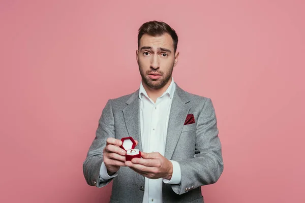 Confused man holding box with proposal ring, isolated on pink — Stock Photo