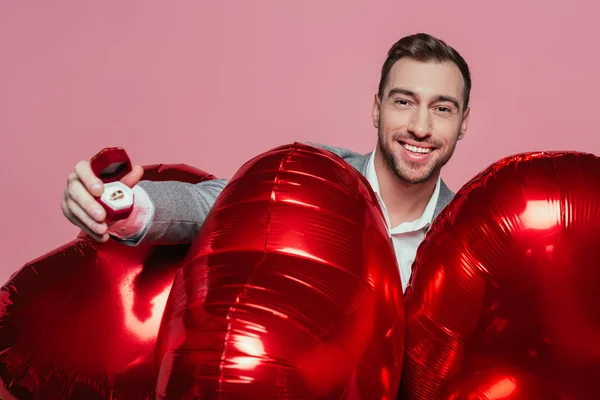 Hombre guapo sosteniendo globos de corazón rojo y anillo de propuesta, aislado en rosa - foto de stock