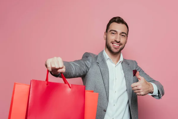 Uomo sorridente tenendo borse della spesa e mostrando pollice in alto, isolato su rosa — Foto stock