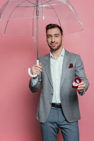 Homem feliz segurando anel proposta e guarda-chuva, isolado em rosa — Fotografia de Stock