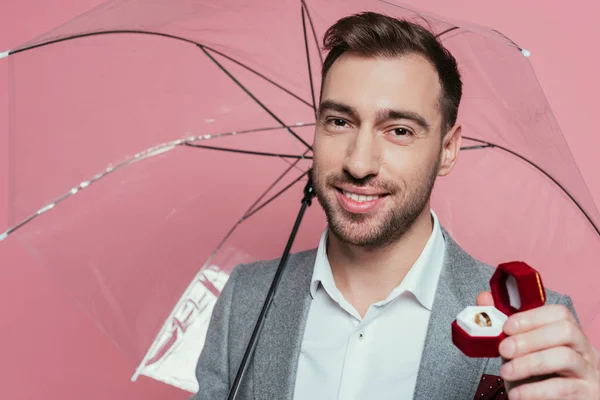 Cheerful man holding proposal ring and umbrella, isolated on pink — Stock Photo