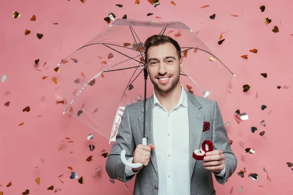 Guapo sonriente hombre sosteniendo anillo de propuesta y paraguas en rosa con corazones confeti - foto de stock