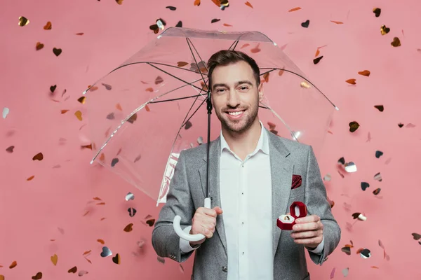 Guapo sonriente hombre sosteniendo anillo de propuesta y paraguas en rosa con corazones confeti — Stock Photo