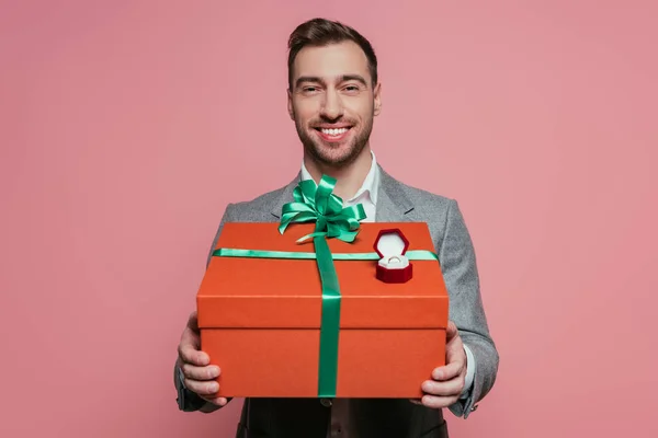 Uomo sorridente in possesso di grande regalo e anello proposta, isolato su rosa — Foto stock