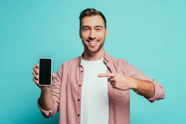 Улыбающийся мужчина указывает на смартфон с пустым экраном, изолированный на синий — стоковое фото