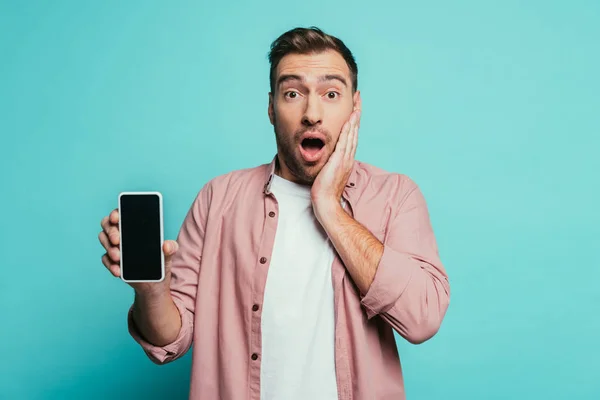 Hombre sorprendido mostrando teléfono inteligente con pantalla en blanco, aislado en azul - foto de stock