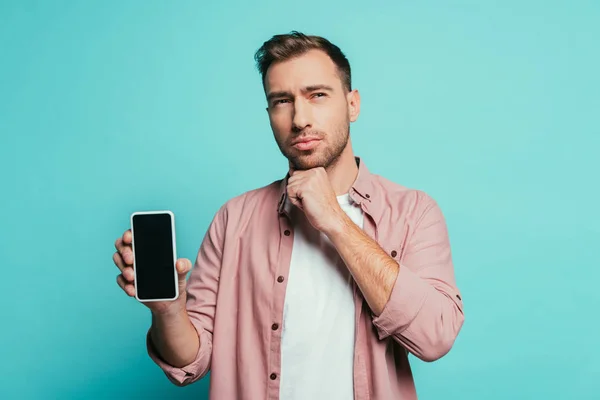 Hombre reflexivo mostrando teléfono inteligente con pantalla en blanco, aislado en azul - foto de stock