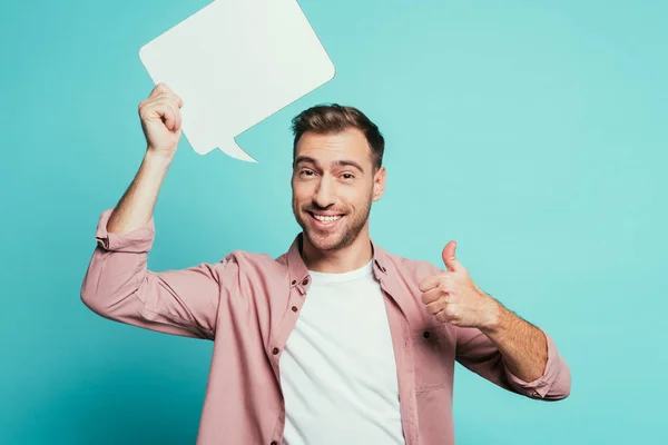 Hombre alegre sosteniendo la burbuja del habla vacía y mostrando el pulgar hacia arriba, aislado en azul — Stock Photo