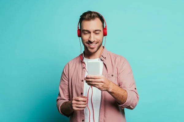 Hombre barbudo feliz escuchando música con auriculares y teléfono inteligente, aislado en azul - foto de stock