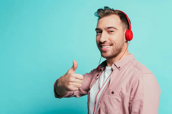 Uomo allegro ascoltare musica con le cuffie e mostrando pollice in alto, isolato su blu — Foto stock