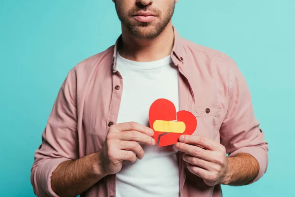 Cropped view of upset man holding broken heart with plaster, isolated on blue — Stock Photo