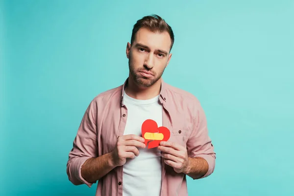 Hombre molesto sosteniendo el corazón roto con yeso, aislado en azul - foto de stock