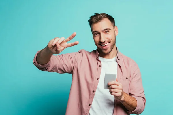 Happy man using smartphone and showing peace symbol, isolated on blue — Stock Photo