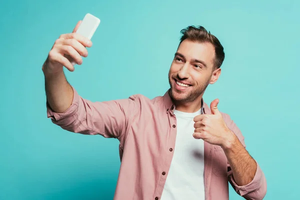 Lächelnder Mann zeigt Daumen hoch und macht Selfie auf Smartphone, isoliert auf blauem Grund — Stockfoto