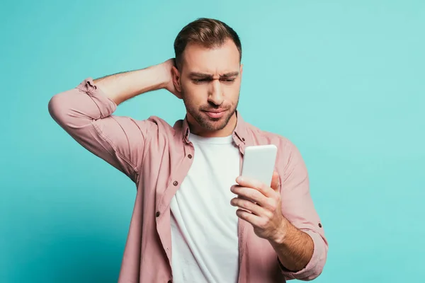 Homem bonito confuso usando smartphone, isolado no azul — Fotografia de Stock