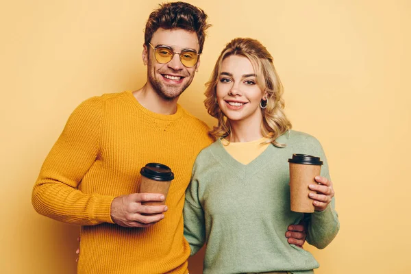 Couple joyeux regardant la caméra tout en tenant du café pour aller sur fond jaune — Photo de stock