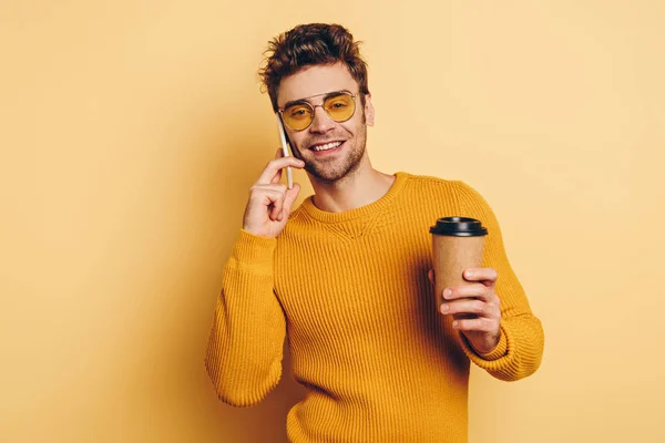 Sonriente hombre hablando en el teléfono inteligente mientras sostiene el café para ir sobre fondo amarillo - foto de stock