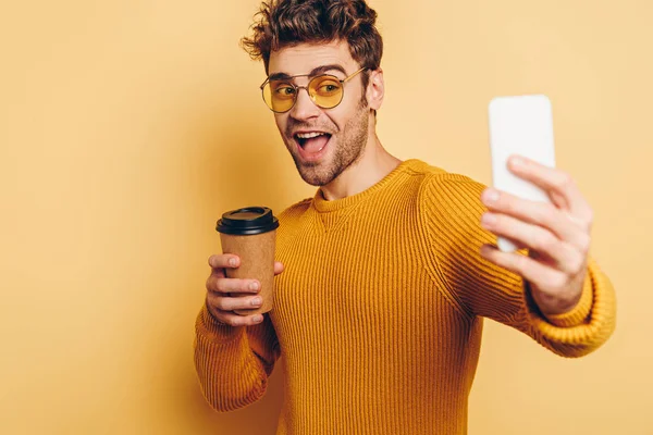 Homem alegre tomando selfie com smartphone enquanto segurando café para ir no fundo amarelo — Fotografia de Stock